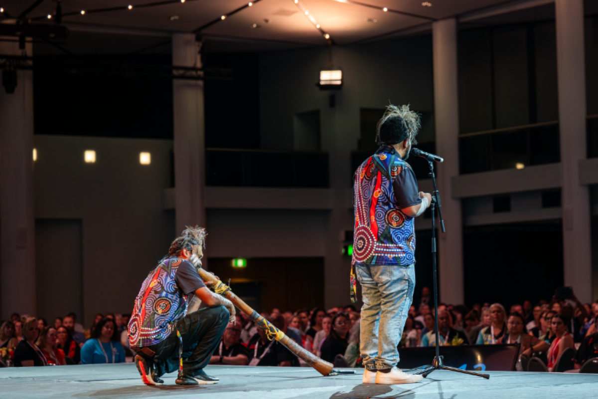 Two Aboriginal performers on stage at a large conference, both wearing traditional dot-print shirts — one speaking at a microphone while the other plays a didgeridoo, with a seated audience visible in the background.
